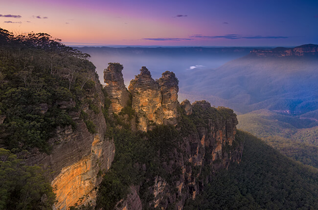 Photo of the three sisters in the Blue Mountains at dawn, with rippling hills covered in vegetation in background